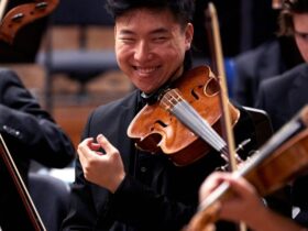 A violinist sitting amongst other musicians, smiling, and wearing a black suit.