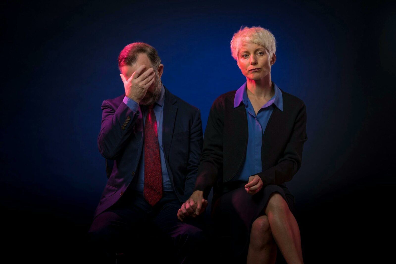 A man and a woman sit side by side against a blue background. The man has his hand over his face.